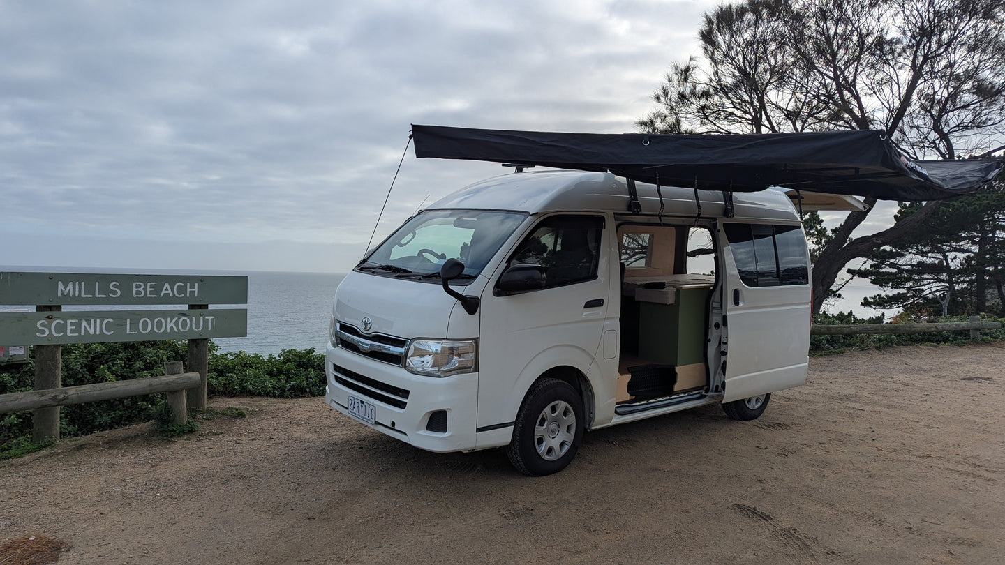A  Toyota HiAce fitted with an OPENROAD car awning, providing full shade coverage beside the vehicle during an outdoor setup.