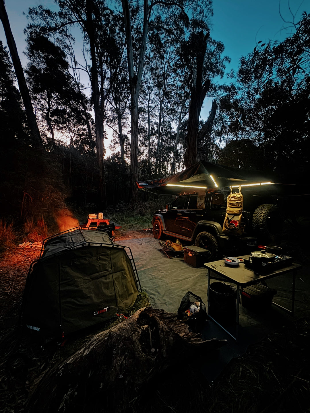 Ineos Grenadier using an OPENROAD 4WD awning at night, illuminated at a forest campsite with tents nearby.