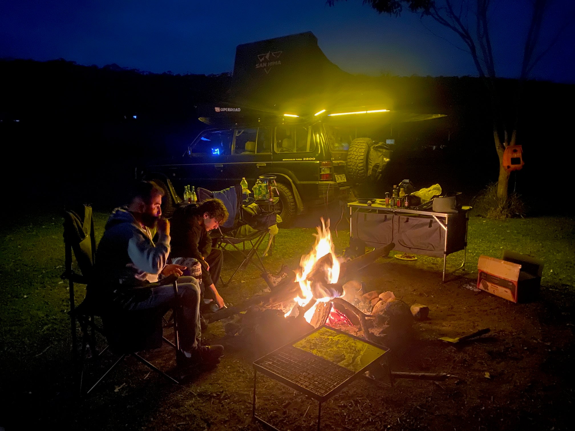 Two campers sitting by a fire in front of a Mitsubishi Pajero with an OPENROAD 270 awning glowing at night like a car awning tent